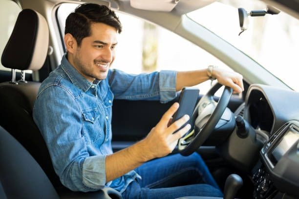 Man sitting in a car's driver's seat, smiling and holding a smartphone while looking at it.